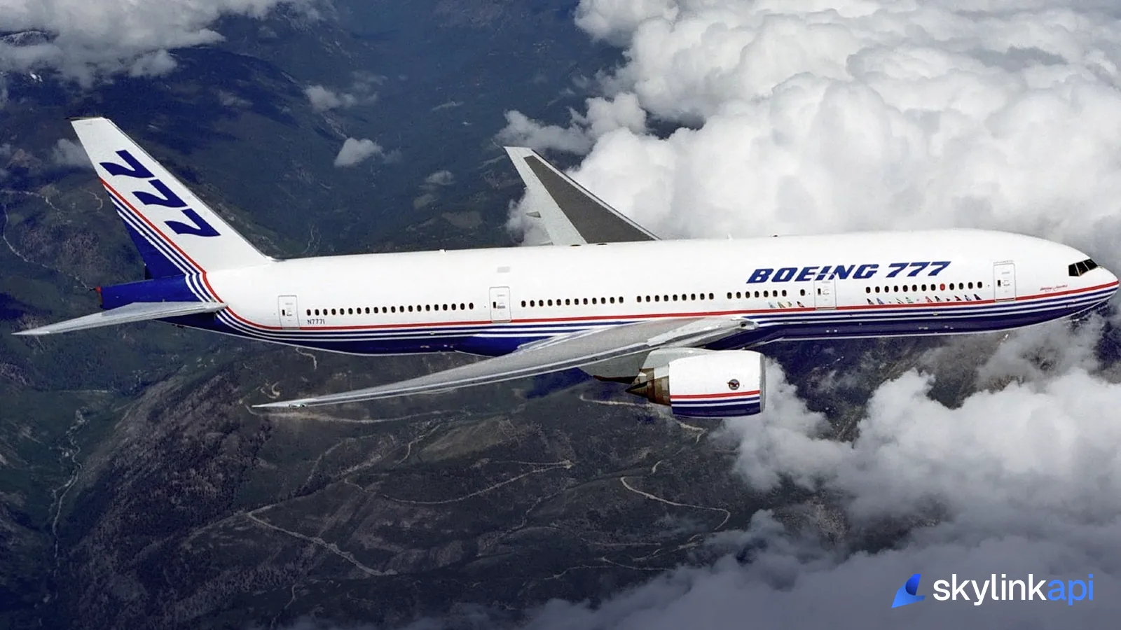 A Boeing 777 in cruise above broken cloud over mountainous terrain